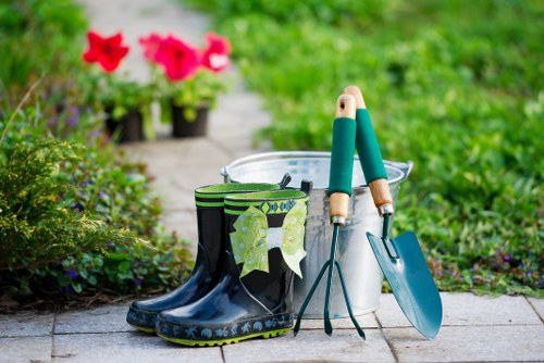 The image depicts a garden scene featuring a pair of black waterproof boots with a green and white leaf pattern near the top, positioned on a paved pathway. Resting against the boots is a pair of gardening tools with wooden handles and metal heads, including a small hand rake and a trowel. To the right, there is a transparent plastic bucket, partially filled with gardening gloves and other tools. In the background, vibrant pink and red flowers in pots are visible, with lush greenery and a blurred garden bed further behind. The scene is set outdoors on a bright, overcast day, with natural light illuminating the various gardening elements, suggesting an environment suitable for maintenance, planting, and lawncare activities typical of professional gardening services in Kentish Town.